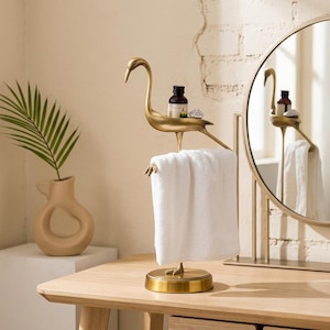May include: A gold-toned metal towel holder shaped like a standing crane with a flat back platform. A small glass bottle with a dark cap sits on the crane's back, and a white rectangular hand towel hangs from the bird's legs over a round base.