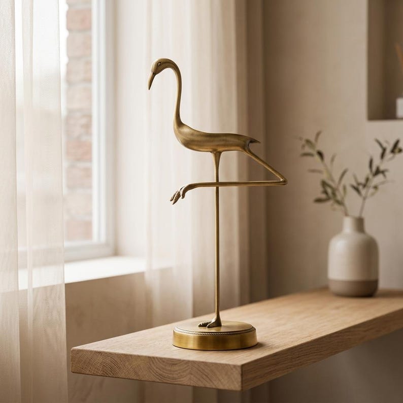 May include: A brass-colored metal sculpture of a crane standing on one leg. The bird figure features a long neck, a pointed beak, and a slender body, mounted on a circular base with a textured edge. The sculpture has a smooth, matte finish.
