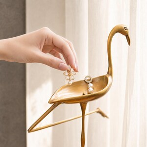 May include: A gold-toned metal bird-shaped jewelry dish with a long neck and slender legs. The dish holds a pair of earrings, one shaped like a flower with white petals and a central pearl, and the other featuring a circular crystal and pearl drop.