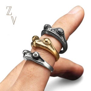 May include: Three metal rings shaped like frogs, stacked on a finger. The top ring is dark grey, the middle ring is gold-toned, and the bottom ring is silver-toned. Each ring features a detailed frog head design with prominent eyes. The letters ZV appear.