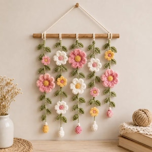 May include: A wall hanging featuring five vertical strands of crocheted green vines with pink, white, and yellow flowers suspended from a horizontal wooden dowel. The piece is held by a white cord attached to the ends of the wooden rod for display.