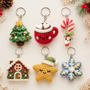 Op de afbeelding: Zes gehaakte sleutelhangers met kerstthema: een groene kerstboom met een gele ster, een rode mok warme chocolademelk met hartvormige marshmallows, een rood-witte zuurstok, een bruin peperkoekhuisje, een gele ster met een groene muts en een blauwe sneeuwvlok.