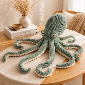 Könnte beinhalten: Ein salbeigrünes, gehäkeltes Oktopus-Stofftier mit einem großen, abgerundeten Kopf, zwei schläfrigen Augen mit dunklen Lidern und acht langen, geringelten Tentakeln. Die Unterseite jedes Tentakels ist mit kleinen, cremefarbenen Häkel-Saugnäpfen besetzt.