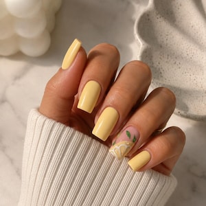 Peut inclure: Un ensemble d'ongles artificiels de forme carrée avec une finition jaune pastel brillante. Un ongle est orné d'un motif peint à la main représentant une tranche de citron avec des feuilles vertes sur une base transparente, les autres sont unis.