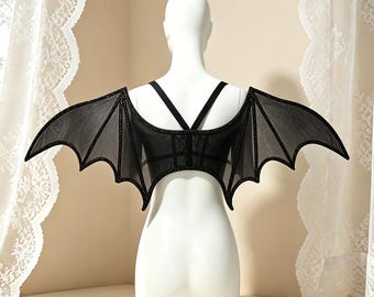 May include: A black garment featuring large, structured bat-like wings extending from the back. The wings are made of a sheer, dark mesh material with defined, scalloped edges and thin straps that cross over the upper back area of the torso.