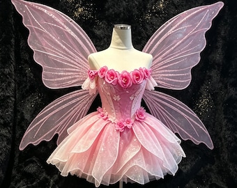 May include: A pink fairy costume featuring large, glittery, translucent wings and a dress adorned with pink fabric roses. The bodice has an off-the-shoulder neckline with floral accents, and the skirt consists of layered, shimmering tulle fabric.