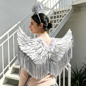 May include: A pair of silver feathered wings with long metallic silver fringe trim attached to a light pink jumper. A matching silver headband features a large, round, metallic silver pom-pom topper that sits above the head.