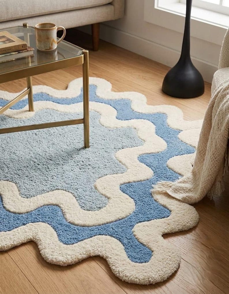 Op de afbeelding: Een getuft karpet met een golvende, organische vorm en concentrische lagen in cr&egrave;me en lichtblauw. Het kleed heeft een dik, getextureerd oppervlak en volgt een herhalend geschulpt patroon dat vanuit het midden naar buiten straalt.