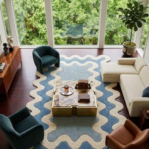 Op de afbeelding: Een groot vloerkleed met een golvende, geschulpte rand met concentrische banden in cr&egrave;me en blauw. Het kleed ligt centraal in een kamer met een vierkante salontafel met een marmeren blad en gouden voet, twee blauwgroene fauteuils en een cr&egrave;mekleurige bank.