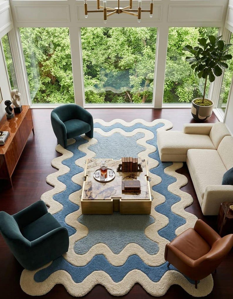 Op de afbeelding: Een groot vloerkleed met een golvende, geschulpte rand met concentrische banden in cr&egrave;me en blauw. Het kleed ligt centraal in een kamer met een vierkante salontafel met een marmeren blad en gouden voet, twee blauwgroene fauteuils en een cr&egrave;mekleurige bank.