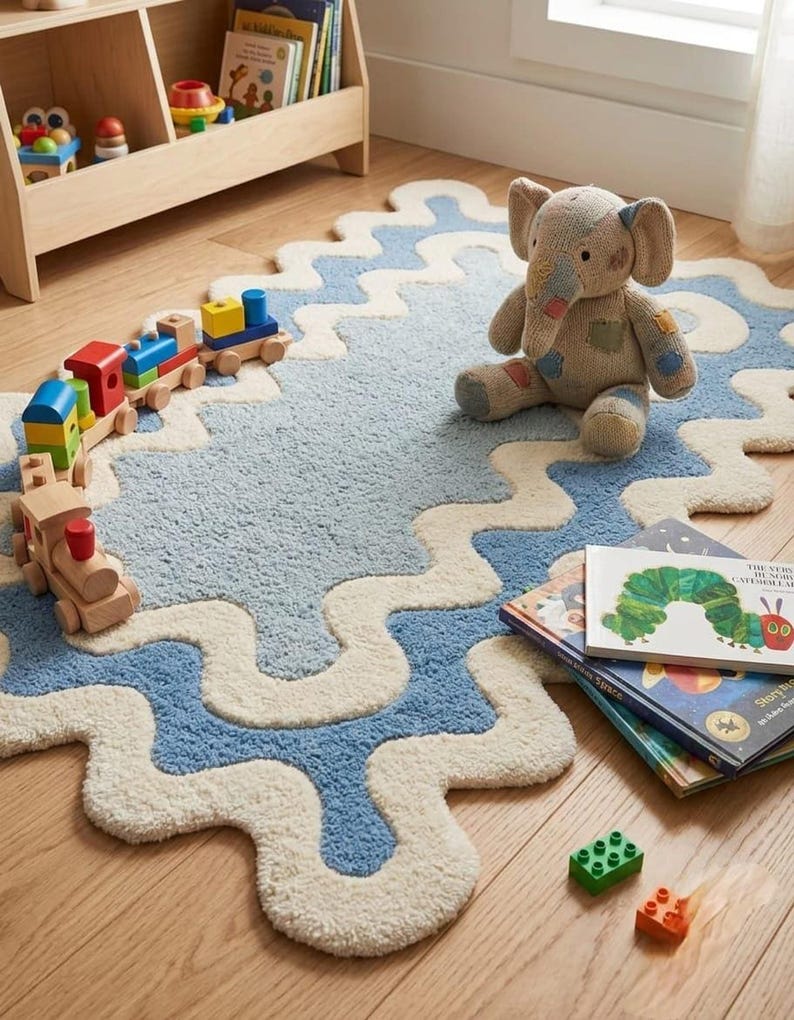Op de afbeelding: Een rechthoekig vloerkleed met een golvende, geschulpte rand in cr&egrave;me en een getextureerd lichtblauw midden. Het kleed is getuft met een zachte, pluizige pool en heeft een opvallend golvend randpatroon dat een organische vorm cre&euml;ert.