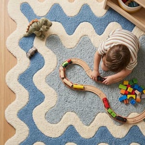 Op de afbeelding: Een rechthoekig vloerkleed met een golvend, geschulpt randpatroon in afwisselende banen van cr&egrave;me en blauw. Het kleed heeft een getextureerd, pluizig oppervlak en is versierd met een houten speelgoedtrein, een kleine groene dinosaurus en blokken.