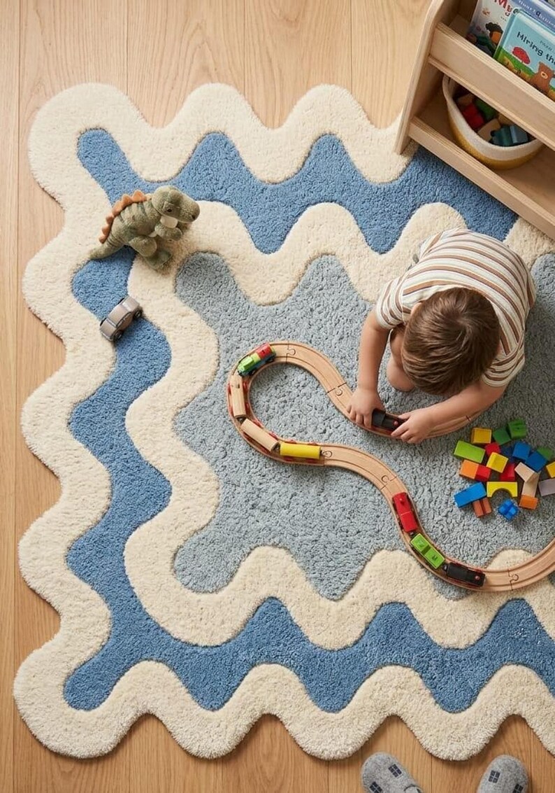 Op de afbeelding: Een rechthoekig vloerkleed met een golvend, geschulpt randpatroon in afwisselende banen van cr&egrave;me en blauw. Het kleed heeft een getextureerd, pluizig oppervlak en is versierd met een houten speelgoedtrein, een kleine groene dinosaurus en blokken.