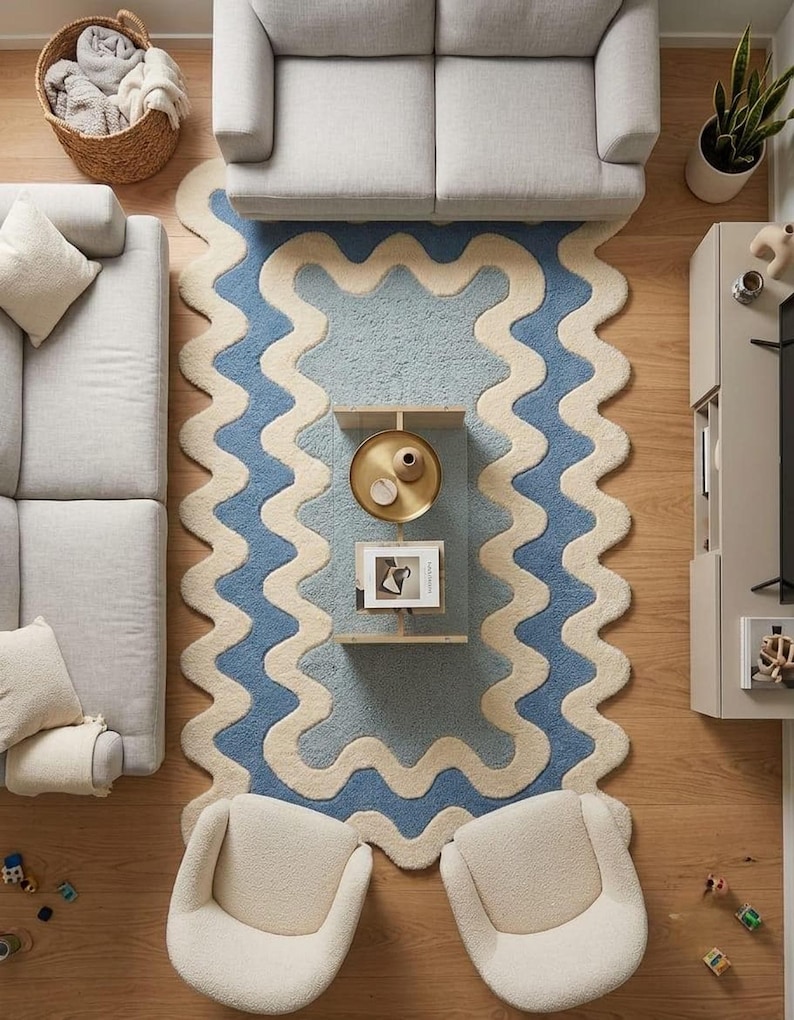 Op de afbeelding: Een rechthoekig vloerkleed met een golvend, geschulpt randontwerp in afwisselende banen van cr&egrave;me en blauw. Het kleed heeft een getextureerd, getuft oppervlak en is centraal geplaatst in een woonkamer tussen twee lichtgrijze banken en stoelen.