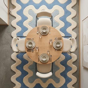 Op de afbeelding: Een ronde eettafel van licht hout met vier cr&egrave;mekleurige stoelen, geplaatst op een groot rechthoekig vloerkleed met een golvend, geschulpt randpatroon in afwisselende banen van cr&egrave;me en blauw. Vier couverts met borden en bestek.