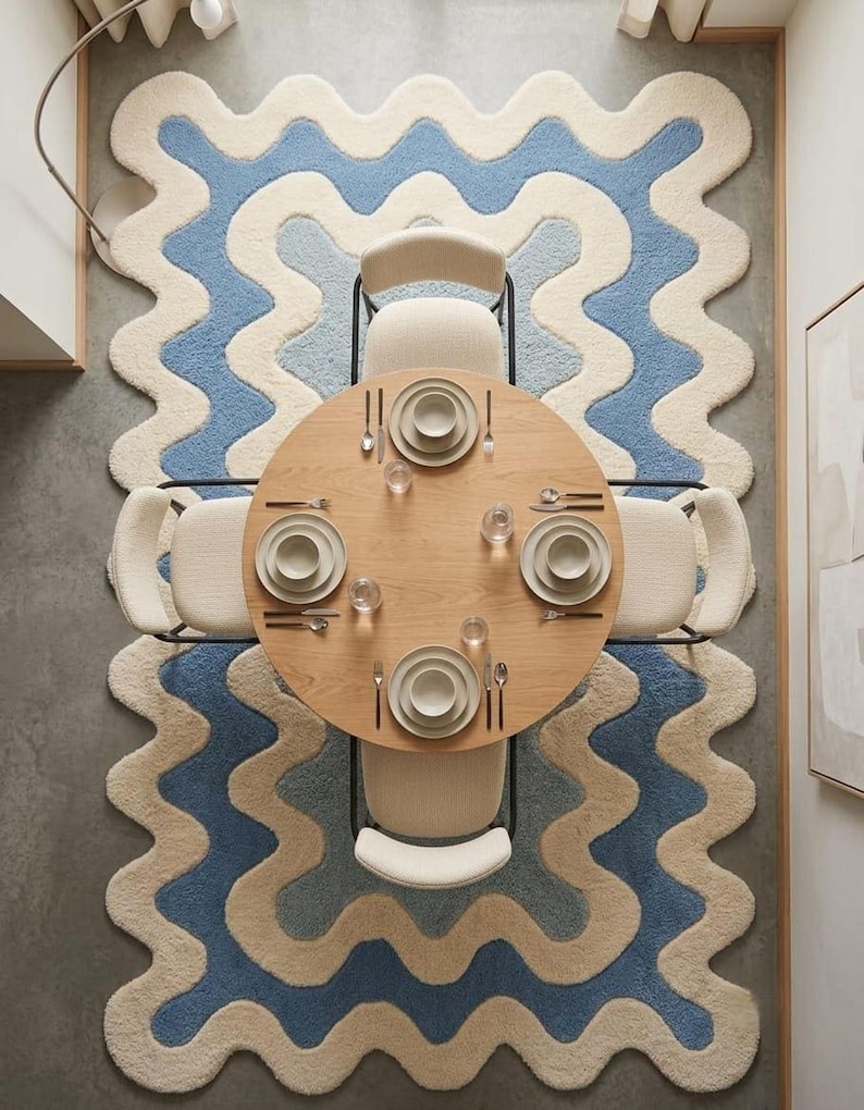 Op de afbeelding: Een ronde eettafel van licht hout met vier cr&egrave;mekleurige stoelen, geplaatst op een groot rechthoekig vloerkleed met een golvend, geschulpt randpatroon in afwisselende banen van cr&egrave;me en blauw. Vier couverts met borden en bestek.