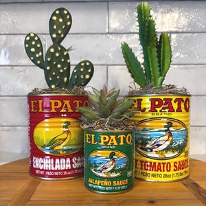 May include: Three decorative planters made from repurposed El Pato brand sauce cans. The cans are painted red, green, and yellow, and feature images of a duck. Each can holds a different type of succulent or cactus.