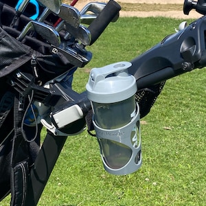 May include: A light gray plastic water bottle with a flip-top lid held in a matching gray silicone cage attached to a black golf cart frame. The bottle features the text Range Bange printed in white on the side of the silicone sleeve.