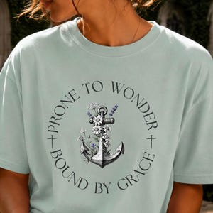May include: A light green short-sleeve t-shirt featuring a graphic of a ship anchor entwined with white daisies and purple flowers. The text "PRONE TO WONDER" and "BOUND BY GRACE" is printed in a circular arrangement around the anchor design.