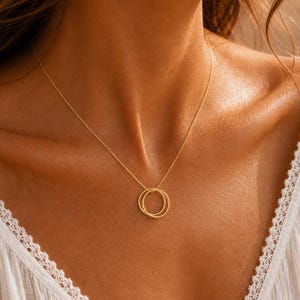May include: A gold-toned necklace featuring a delicate chain and a pendant composed of two interlocking circular rings. The necklace rests against a white garment with a lace-trimmed neckline, highlighting the metallic finish of the jewellery.