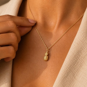 May include: A gold-toned necklace featuring a small, textured pineapple pendant suspended from a delicate chain. The pendant displays a detailed, cross-hatched surface pattern and a leafy top, reflecting light with a polished, metallic finish.