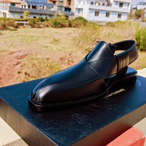 May include: A black leather sandal with a pointed toe, a closed front, and an open heel design featuring a buckled strap. The sandal is displayed on a black rectangular box with the text "Luxury" visible on the surface of the box.