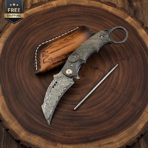 May include: A folding karambit knife with a patterned Damascus steel blade, a dark textured handle with a finger ring, a copper-coloured pivot screw, a tan leather sheath with decorative stitching, and a thin metal sharpening rod.