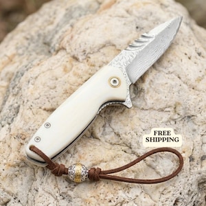 May include: A folding pocket knife with a cream-colored handle and a patterned Damascus steel blade. The handle features a gold-toned pivot screw and a brown leather lanyard with a decorative silver and gold-toned metal bead attached.