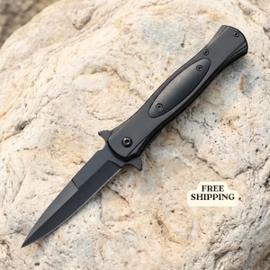 Könnte beinhalten: Ein mattschwarzes Klappmesser mit spitzer Klinge und einem schlanken Griff, der eine mittige Einlage und sichtbare Schrauben aufweist. Das Messer ist diagonal ausgerichtet und zeigt seine scharfe Klinge und das Gelenk.