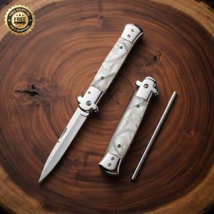 May include: A set consisting of one open stiletto-style folding knife and one closed stiletto-style folding knife, both featuring polished silver metal bolsters and white pearlescent handles, accompanied by a thin, straight silver metal rod.