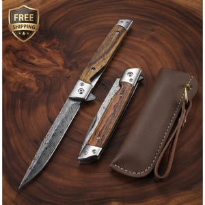 Può includere: Due coltelli a serramanico con lame in acciaio damasco e manici in legno, dotati di bolster e rivetti in metallo color argento. Un coltello è completamente aperto, mentre l'altro è chiuso accanto a un fodero in pelle marrone con cinturino.