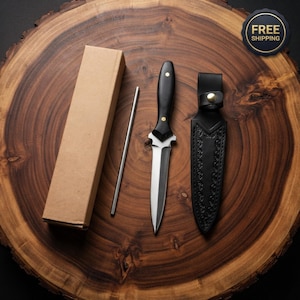 May include: A double-edged dagger with a black handle and two brass rivets, a matching black leather sheath with embossed scrollwork and a brass snap closure, a thin metal rod, and a rectangular cardboard box arranged on a wooden surface.