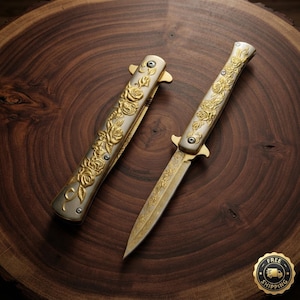 May include: A folding knife with a silver and gold handle featuring an embossed rose pattern, shown alongside its matching silver and gold sheath. The blade is partially extended and displays an intricate, etched floral design along its length.