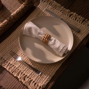 May include: A beige ceramic plate holds a folded off-white cloth napkin secured with a woven tan rattan napkin ring. A silver fork rests on the left side of a beige crocheted placemat with fringed edges, and a silver knife lies on the right side.