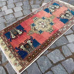 May include: A rectangular woven rug featuring a vibrant coral-red field with a central beige geometric medallion. The design includes symmetrical blue and dark brown motifs at the ends, framed by a patterned border with small fringe at both edges.