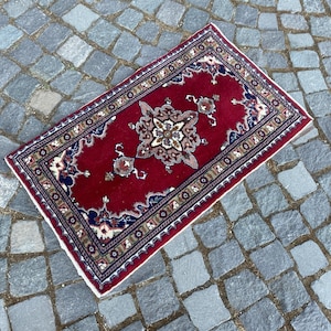 May include: A rectangular area rug featuring a deep red field with a central ornate floral medallion in cream, brown, and blue. The rug is framed by a multi-layered border with intricate geometric and floral patterns in navy, cream, and tan tones.