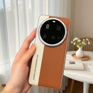 May include: A smartphone case featuring a two-tone design with a white left side and a textured brown right side. The back includes a large circular camera module with the text LEICA, VARIABLE APERTURE, and CAMERA-LIKE PHONESHELL printed on it.