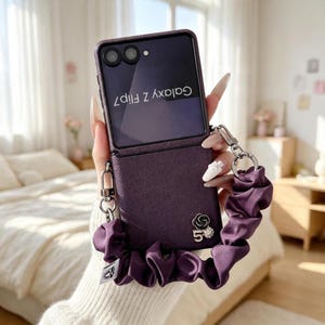 Könnte beinhalten: Eine dunkelviolette Galaxy Z Flip7 Smartphone-Hülle mit strukturierter Oberfläche. Die Hülle verfügt über eine passende, geraffte Stoff-Handschlaufe, die mit silbernen Metallclips befestigt ist, sowie einen kleinen silberfarbenen Blumen- und Fünfer-Anhänger.