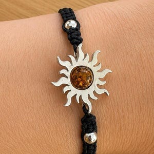 Puede incluir: Una pulsera de cordón negro con un dije de sol plateado y una piedra central de color ámbar. La pulsera incluye dos pequeñas cuentas plateadas en el cordón, una colocada encima y otra debajo del dije de sol, aseguradas con nudos.