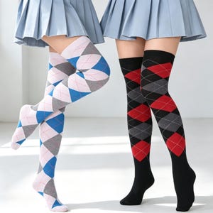 Puede incluir: Dos pares de calcetines por encima de la rodilla usados con faldas plisadas azul claro. Un par presenta un patrón de rombos en rosa claro, azul y gris. El segundo par es negro con un patrón de rombos rojo y gris. Ambos tienen líneas blancas.