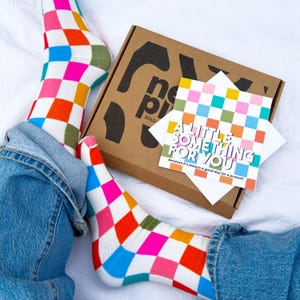 May include: A pair of socks featuring a multicoloured checkerboard pattern in pink, orange, blue, and green, paired with a brown cardboard gift box and a square greeting card that reads A LITTLE SOMETHING FOR YOU because it's always a good day for a present.