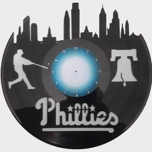 May include: Black vinyl record clock featuring a Philadelphia skyline silhouette, a baseball player, and the Liberty Bell. The clock face has a blue gradient. The word "Phillies" is displayed in white script.