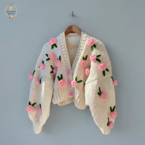 Könnte beinhalten: Eine cremefarbene Strickjacke aus grobem Strick mit langen Ärmeln und V-Ausschnitt. Das Kleidungsstück ist mit dreidimensionalen hellrosa Garnrosen und grünen Blättern verziert, die über die Vorderseite und Ärmel verteilt sind, und hat drei hellrosa Knöpfe.