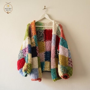 Puede incluir: Un cárdigan de punto grueso con un diseño de parches de cuadrados multicolores en tonos rojo, azul, verde, amarillo, rosa y blanco. La prenda tiene mangas largas, un corte holgado y se muestra en una percha blanca contra una pared lisa.