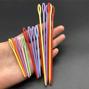 May include: A collection of plastic sewing needles in various bright colours including red, yellow, blue, green, orange, purple, and pink. Each needle features a large, elongated eye at the top and a pointed tip at the bottom, arranged in a fan shape.