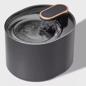 Può includere: Una fontanella per animali domestici di forma ovale, di colore nero con una finitura opaca liscia. La superficie superiore presenta un pannello di controllo o sensore rettangolare arrotondato con un bordo color rame, con acqua chiara.