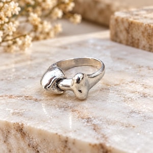 May include: A silver-toned metal ring featuring a sculptural design that resembles a bone or organic shape. The ring has a smooth, polished finish with a curved band that tapers toward the focal point, creating a continuous, fluid silhouette.