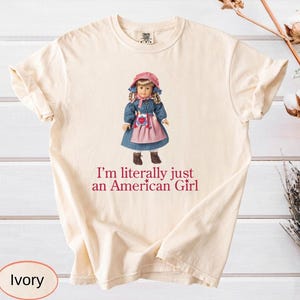 May include: An ivory short-sleeved t-shirt featuring a graphic of a doll wearing a blue dress, a pink apron, and a pink bonnet. Below the doll, the text reads, I'm literally just an American Girl, printed in a dark red, serif font.