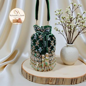 Può includere: Una borsa a sacchetto in velluto verde scuro con coulisse, decorata con intricate perline nei toni dell'oro, dell'argento e del verde chiaro. La borsa presenta un manico in velluto verde abbinato e una frangia decorativa di perline pendenti.