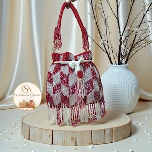 Könnte beinhalten: Eine Beuteltasche mit Kordelzug, die mit einem geometrischen Muster aus roten und silbernen Perlen bedeckt ist. Die Tasche verfügt über einen einzelnen Perlenhenkel, einen weißen Kordelzug mit einem runden Perlenakzent und lange Fransen.